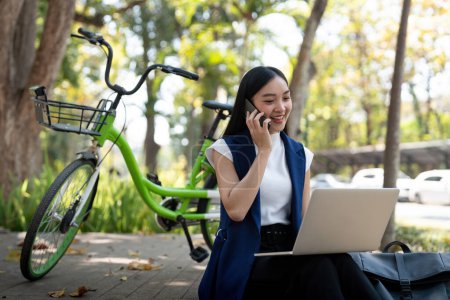 Asiatische Geschäftsfrau mit Fahrrad, Laptop und Smartphone vor dem Bürogebäude. Frauen, die mit dem Fahrrad zur Arbeit fahren. Umweltfreundliches Fahrzeug, nachhaltiges Lifestylekonzept.