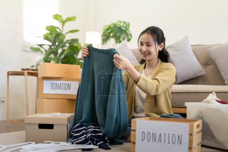 Foto de Mujer asiática sosteniendo caja de donación llena de ropa y ropa selecta. Concepto de donación y reciclaje de ropa. Ayudar a los pobres. - Imagen libre de derechos