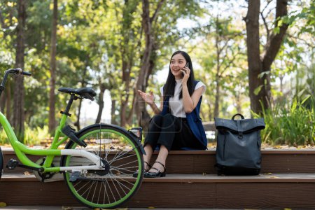 Asian businesswoman with bicycle using smartphone and sitting outside the office building. Woman commuting on bike go to work. Eco friendly vehicle, sustainable lifestyle concept.