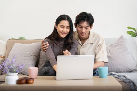 Téléchargez les photos : Heureux couple asiatique à l'aide d'un ordinateur portable assis se détendre sur le canapé faire du commerce électronique shopping en ligne sur le site à la maison. - en image libre de droit