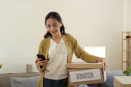 Foto de Concepto de donación. Mujer asiática sosteniendo una caja de donaciones con ropa llena. Mujer sosteniendo ropa donar caja. Ropa en caja para la donación de concepto y reciclaje de reutilización. - Imagen libre de derechos