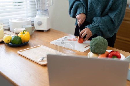 Photo for A woman is chopping plantbased food on a hardwood countertop while following a recipe on a laptop. The gesture of slicing fruit shows her culinary skills - Royalty Free Image