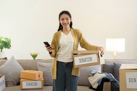 Foto de Concepto de donación. Mujer asiática sosteniendo una caja de donaciones con ropa llena. Mujer sosteniendo ropa donar caja. Ropa en caja para la donación de concepto y reciclaje de reutilización. - Imagen libre de derechos
