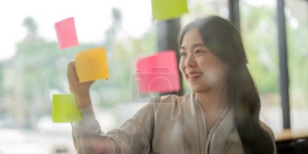 Photo for Smiling businesswoman Asian people thinking and use sticky notes on glass wall in office. - Royalty Free Image