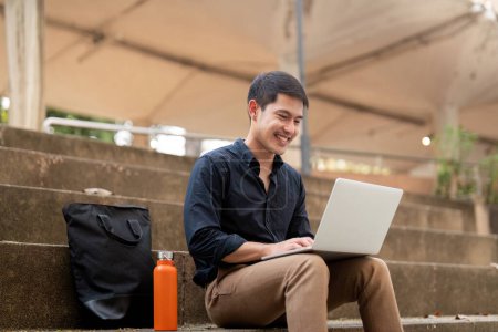Téléchargez les photos : Homme d'affaires asiatique avec tasse écologique réutilisable en utilisant un ordinateur portable et assis à l'extérieur de l'immeuble de bureaux. Concept de mode de vie écologique et durable. - en image libre de droit