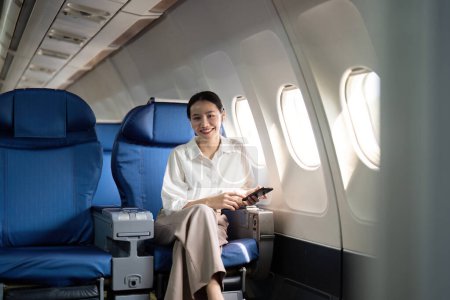 Foto de Viajes y tecnología. Volando en primera clase. Joven pasajera de negocios usando smartphone mientras está sentada en vuelo en avión. - Imagen libre de derechos