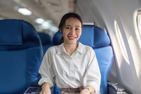 Photo for A woman is sitting on a blue airplane seat with a smile on her face. She is wearing a white shirt and a ponytail - Royalty Free Image