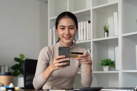 Photo for Happy young asian woman holding credit card using mobile payment at home. Smiling Female customer shopping making purchase on smartphone receiving cash back concept. - Royalty Free Image