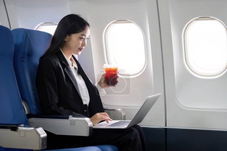 Téléchargez les photos : Asiatique jeune femme utilisant un ordinateur portable tout en buvant du café en première classe dans l'avion pendant le vol, Voyager et Business concept. - en image libre de droit