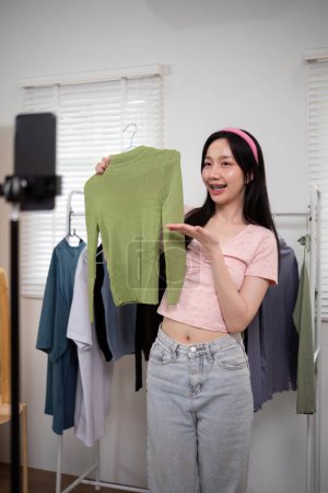 Foto de Mujer asiática venta en línea está utilizando el teléfono inteligente para mientras que la revisión de la colección de camisas y hablar con el cliente a la venta de ropa en línea en las redes sociales. - Imagen libre de derechos