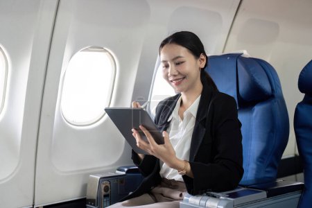 Foto de Sonriente feliz mujer de negocios asiática volando y trabajando en un avión en primera clase, Mujer sentada dentro de un avión usando tableta digital. - Imagen libre de derechos