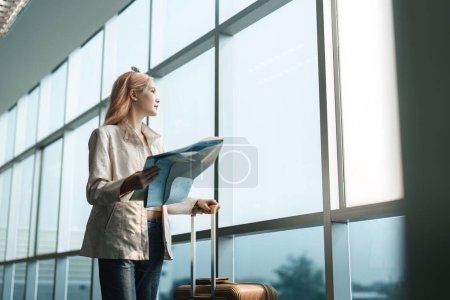 Téléchargez les photos : Heureuse jeune femme asiatique est debout à l'aéroport près de la valise et de la carte de lecture. - en image libre de droit