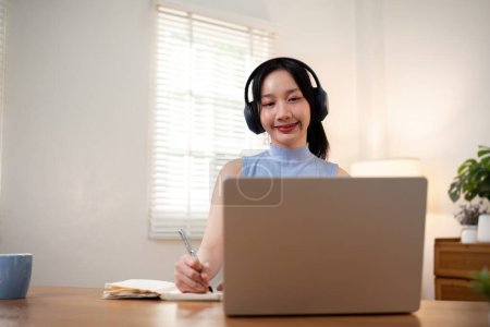 Téléchargez les photos : Heureuse jeune femme asiatique dans les écouteurs et ordinateur portable prendre des notes, fille étudiante parler par vidéoconférence, formation en ligne. - en image libre de droit