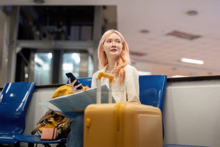 Foto de Feliz joven mujer asiático es sentado en aeropuerto cerca maleta y lectura mapa. - Imagen libre de derechos