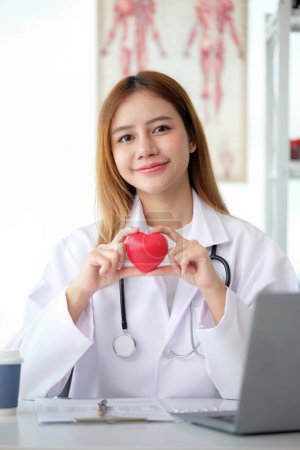 Foto de Feliz joven doctora sosteniendo un objeto rojo en forma de corazón, mirando a la cámara con sonrisa. Practicante positivo, cardiólogo. - Imagen libre de derechos
