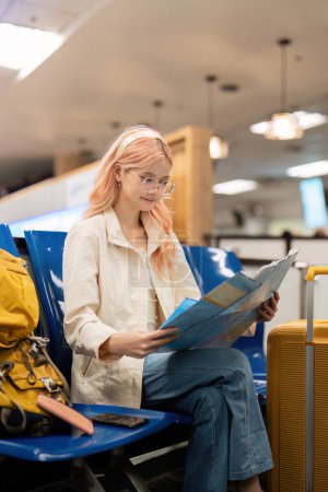 Téléchargez les photos : Heureuse jeune femme asiatique est assis à l'aéroport près de la valise et de la carte de lecture. - en image libre de droit