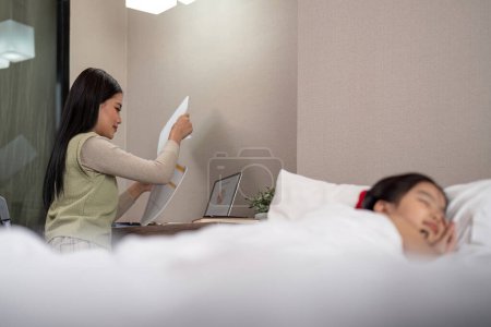Téléchargez les photos : Mère asiatique travaillant sur le lit avec fille endormie à la maison. - en image libre de droit
