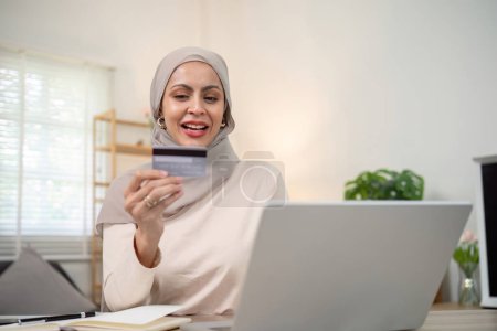 Téléchargez les photos : Jeune femme musulmane heureuse dans le hijab à la maison en utilisant un ordinateur portable shopping en ligne avec carte de crédit tout en étant assis sur le bureau. - en image libre de droit