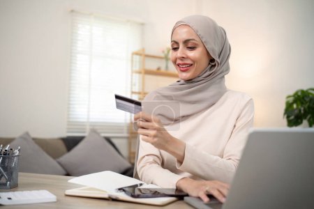 Téléchargez les photos : Jeune femme musulmane heureuse dans le hijab à la maison en utilisant un ordinateur portable shopping en ligne avec carte de crédit tout en étant assis sur le bureau. - en image libre de droit