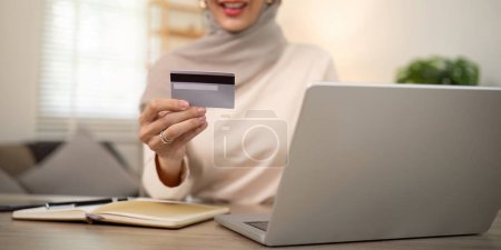 Foto de Joven mujer musulmana feliz en hijab en casa usando el ordenador portátil de compras en línea con tarjeta de crédito mientras está sentado en el escritorio. - Imagen libre de derechos