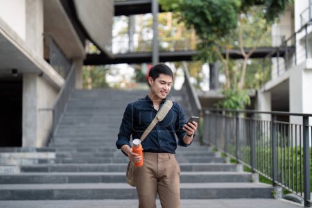 Lächelnder asiatischer Geschäftsmann, der zur Arbeit läuft, während er in der Stadt umweltfreundliche Mehrwegbecher hält.