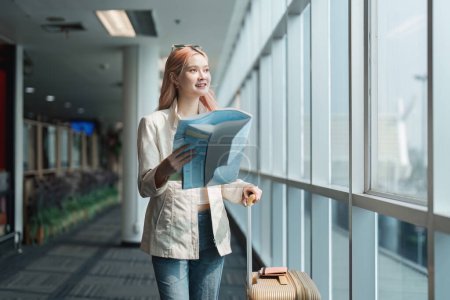 Téléchargez les photos : Heureuse jeune femme asiatique est debout à l'aéroport près de la valise et de la carte de lecture. - en image libre de droit
