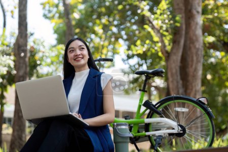 Photo for Asian businesswoman with bicycle using laptop and sitting outside the office building. Woman commuting on bike go to work. Eco friendly vehicle, sustainable lifestyle concept. - Royalty Free Image