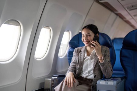 Foto de Asiática joven empresaria exitosa o empresaria en traje formal en un avión sentarse en un asiento de clase ejecutiva y utiliza un teléfono inteligente durante el vuelo. Concepto de Viajes y Negocios. - Imagen libre de derechos