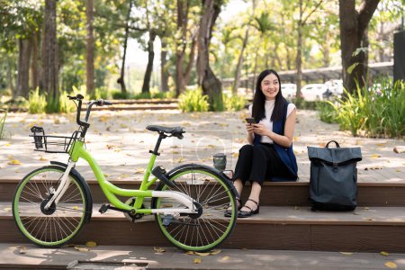 Téléchargez les photos : Femme d'affaires asiatique avec vélo utilisant un smartphone et assise à l'extérieur de l'immeuble de bureaux. Les femmes qui font la navette à vélo vont au travail. Véhicule écologique, concept de mode de vie durable. - en image libre de droit