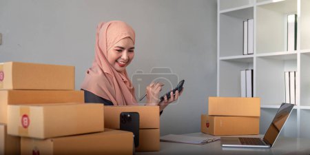 Photo for Muslim women selling online at home with box. Selling online with box to accept order from customer. SME business idea. Parcel delivery. muslim woman working smartphone and laptop at home. - Royalty Free Image