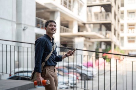 Téléchargez les photos : Souriant asiatique homme d'affaires marchant au travail tout en tenant réutilisable éco-tasse écologique dans la ville. - en image libre de droit