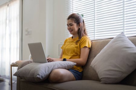 Téléchargez les photos : Jeune femme assise sur le canapé et travaillant sur un projet, regardant un film sur un ordinateur portable se reposer et bavarder heureux avec un ami dans un réseau social à la maison. - en image libre de droit