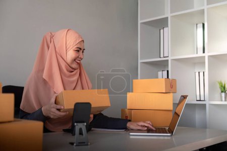 Téléchargez les photos : Femmes musulmanes vendant en ligne à la maison avec boîte. Vente en ligne avec boîte pour accepter la commande du client. Idée d'entreprise PME. Livraison de colis. femme musulmane travailler smartphone et ordinateur portable à la maison. - en image libre de droit