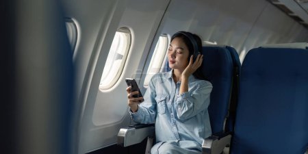 Photo for Asian woman traveler in airplane wearing headset listening music from mobile phone going on a trip vacation travel concept. - Royalty Free Image