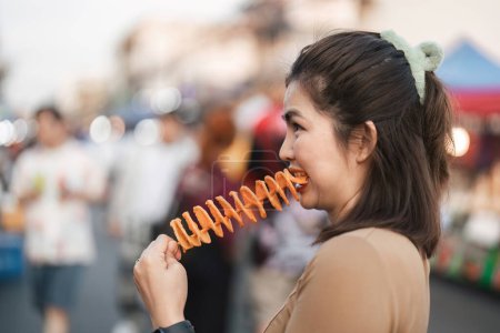 Téléchargez les photos : Heureux jeune asiatique voyageur foodie femme profiter de manger Tornado pommes de terre frites à l'extérieur nuit marché street food. - en image libre de droit