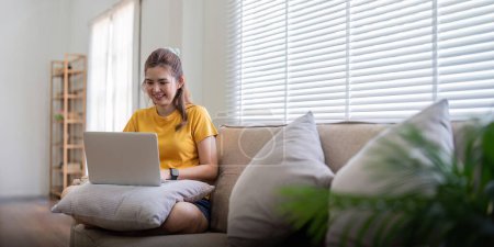Téléchargez les photos : Jeune femme assise sur le canapé et travaillant sur un projet, regardant un film sur un ordinateur portable se reposer et bavarder heureux avec un ami dans un réseau social à la maison. - en image libre de droit