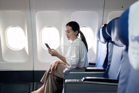 Téléchargez les photos : Une femme est assise dans un avion et regarde son téléphone. Elle sourit et profite de son temps dans l'avion - en image libre de droit