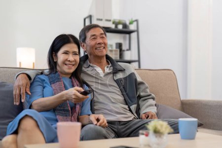 Téléchargez les photos : Asiatique couple aîné profiter de leur télévision préférée. Joyeux couple de retraités regardant la télévision ensemble dans le salon. - en image libre de droit
