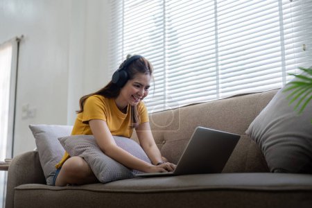 Téléchargez les photos : Femme heureuse portant des écouteurs regardant et écoutant des vidéos sur ordinateur portable et écoutant de la musique assise sur un canapé à la maison. - en image libre de droit