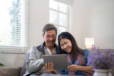 Photo for Happy elderly asian couple using tablet sit on sofa doing ecommerce shopping online on website and buying insurance browsing at home. - Royalty Free Image