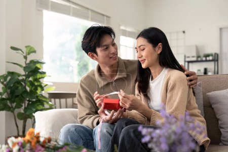 Téléchargez les photos : Romantique jeune couple asiatique embrassant donner cadeau dans le salon à la maison. Tomber amoureux. Concept de Valentin. - en image libre de droit