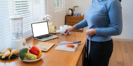 Foto de Alegre mujer gorda cocina en la cocina mostrando portátil maqueta de pantalla blanca en blanco en casa. - Imagen libre de derechos