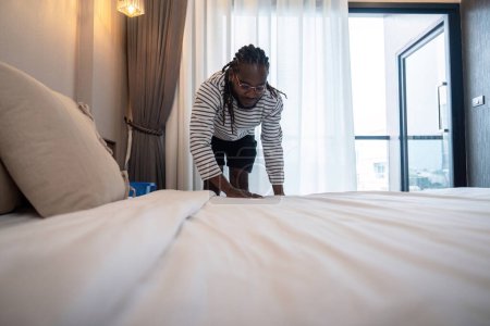 Téléchargez les photos : Jeune homme afro-américain propre faisant le lit. heureux homme noir changer draps à la maison debout dans la chambre. - en image libre de droit
