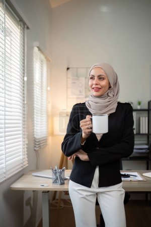 Téléchargez les photos : Femme d'affaires musulmane. femme d'affaires confiante musulmane en hijab debout avec tasse de café du matin au bureau. - en image libre de droit