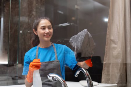 Téléchargez les photos : Femme asiatique nettoyer le verre dans l'appartement. Le personnel de nettoyage maintient la propreté avec une serviette et un détergent pulvérisé. - en image libre de droit