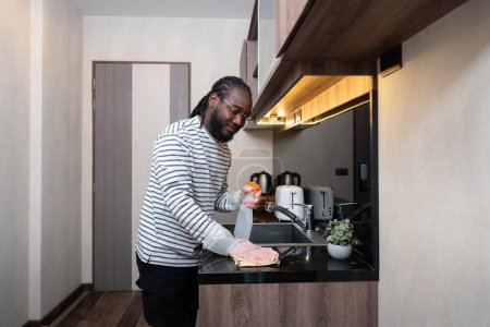 Foto de El hombre afroamericano con guantes de goma limpia el polvo de la mesa en la cocina, las tareas domésticas, la limpieza, el estilo de vida, el hogar. - Imagen libre de derechos