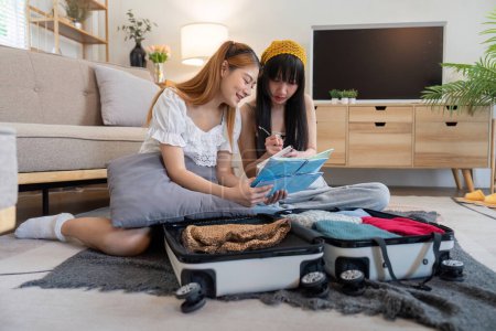 Photo for Young Asian woman packing clothes to the suitcase. Preparation for the summertime vacation. Two women are planning a trip and helping to prepare luggage to travel. - Royalty Free Image