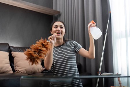 Photo for House cleaning with fun. Happy young asian housewife singing song during cleanup, using feather duster as microphone, enjoying domestic work. Young woman dancing and cleaning in living room. - Royalty Free Image