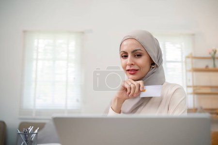 Foto de Joven mujer musulmana feliz en hijab en casa usando el ordenador portátil de compras en línea con tarjeta de crédito mientras está sentado en el escritorio. - Imagen libre de derechos