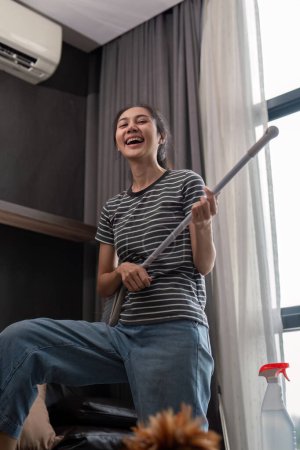 Téléchargez les photos : House cleaning with fun. Happy young asian housewife singing song during cleanup, using mop as guitar, enjoying domestic work. Young woman dancing and cleaning in living room. - en image libre de droit